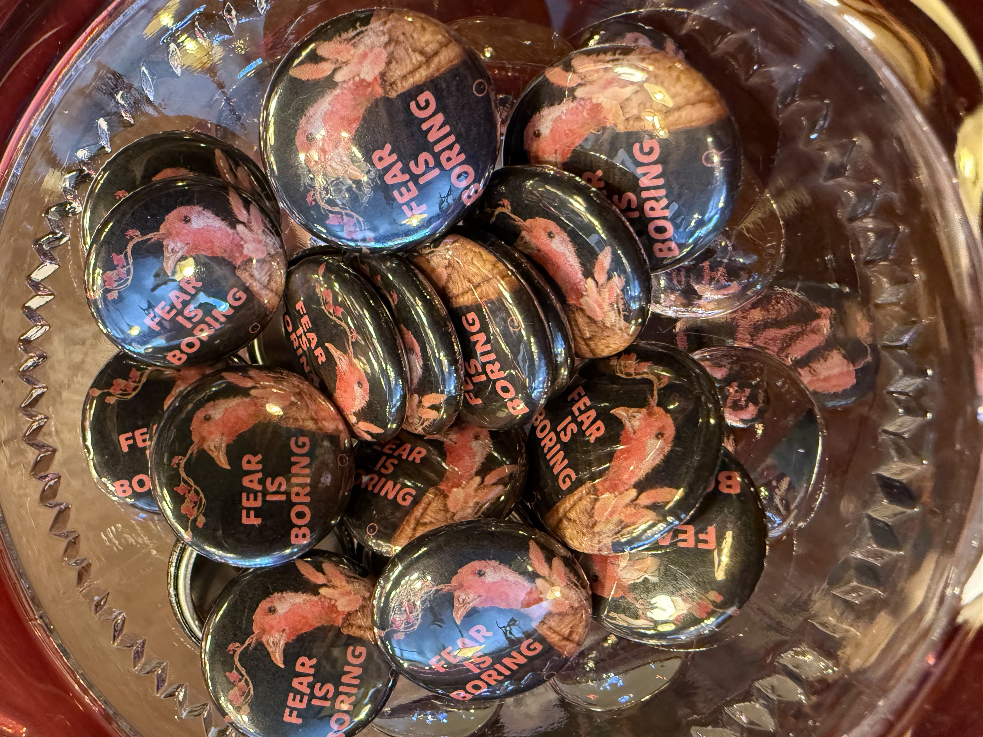 A close-up of pin-back buttons in a glass bowl. Each features Marcellus's portrait on a black background with the words 'Fear is boring' in pink.